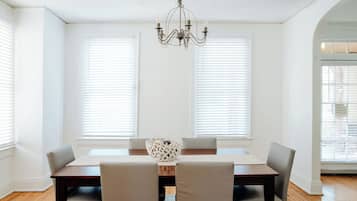 A dining room with a wooden table, four chairs, a chandelier, and large windows with blinds.