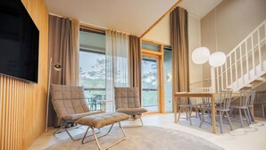 Uma sala de estar moderna com duas poltronas estofadas, uma mesa de jantar com cadeiras, uma TV fixada na parede e grandes janelas com cortinas.
