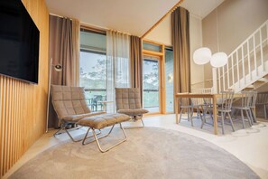 Una sala moderna con dos sillones mullidos, una mesa de comedor con sillas, un televisor montado en la pared y ventanas amplias con cortinas.