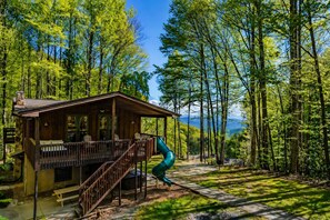 Eine Hütte mit einer Terrasse, einer Rutsche und Blick auf einen bewaldeten Bereich.