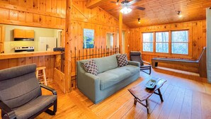 L’intérieur d’une cabane en rondins comprend un coin cuisine, un canapé, un fauteuil et une fenêtre donnant sur la forêt.