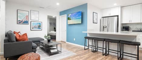A modern living room with a grey sofa, a coffee table, and a small orange ottoman. There is a kitchen area with white cabinets and a stainless steel refrigerator.