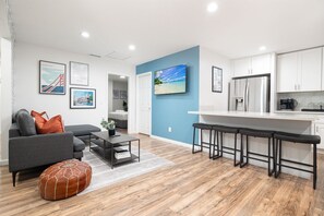 Een moderne woonkamer met een grijze bank, een salontafel en een klein oranje pouf. Er is een keuken met witte kasten en een roestvrijstalen koelkast.