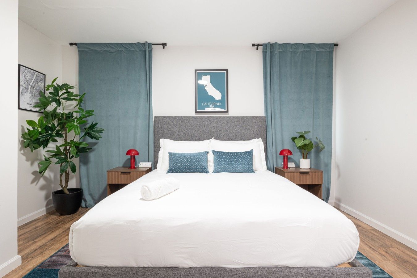 A neatly made bed with white linens, two bedside tables with lamps, a framed map of California, and a potted plant on the left.