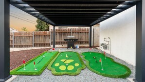 A covered patio area with a miniature golf setup, a barbecue grill, and a wooden fence in the background.
