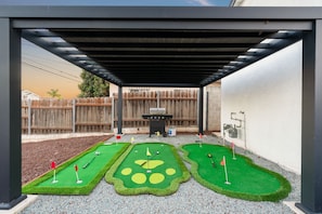 A covered patio area with a miniature golf setup, a barbecue grill, and a wooden fence in the background. - Triple Hot Tub Heaven: Fire Pit & Gameroom (San Diego)