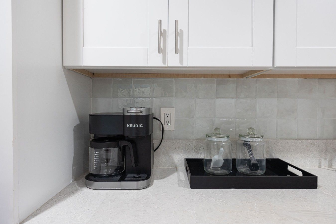 Pieni keittiönurkkaus, jossa on Keurig-kahvinkeitin, kaksi lasipurkkia ja valkoiset kaapit.