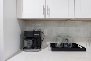 Una kitchenette con una cafetera Keurig, dos frascos de vidrio y armarios blancos.