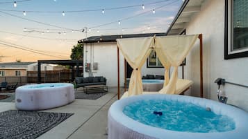 A backyard with two hot tubs, a covered seating area, and string lights.