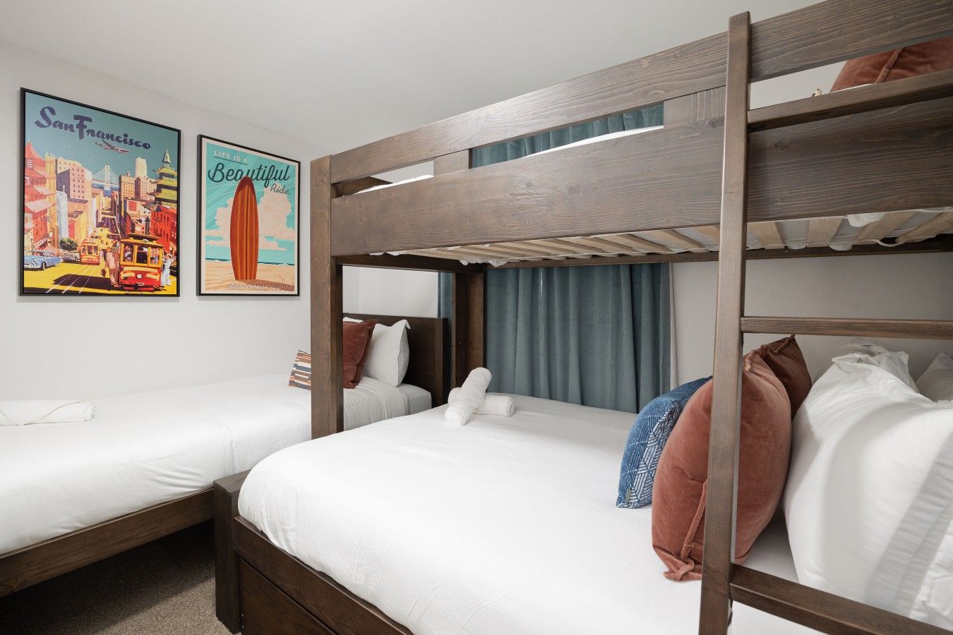 A hotel room with two single beds and a bunk bed, featuring a cityscape and beach-themed wall art.
