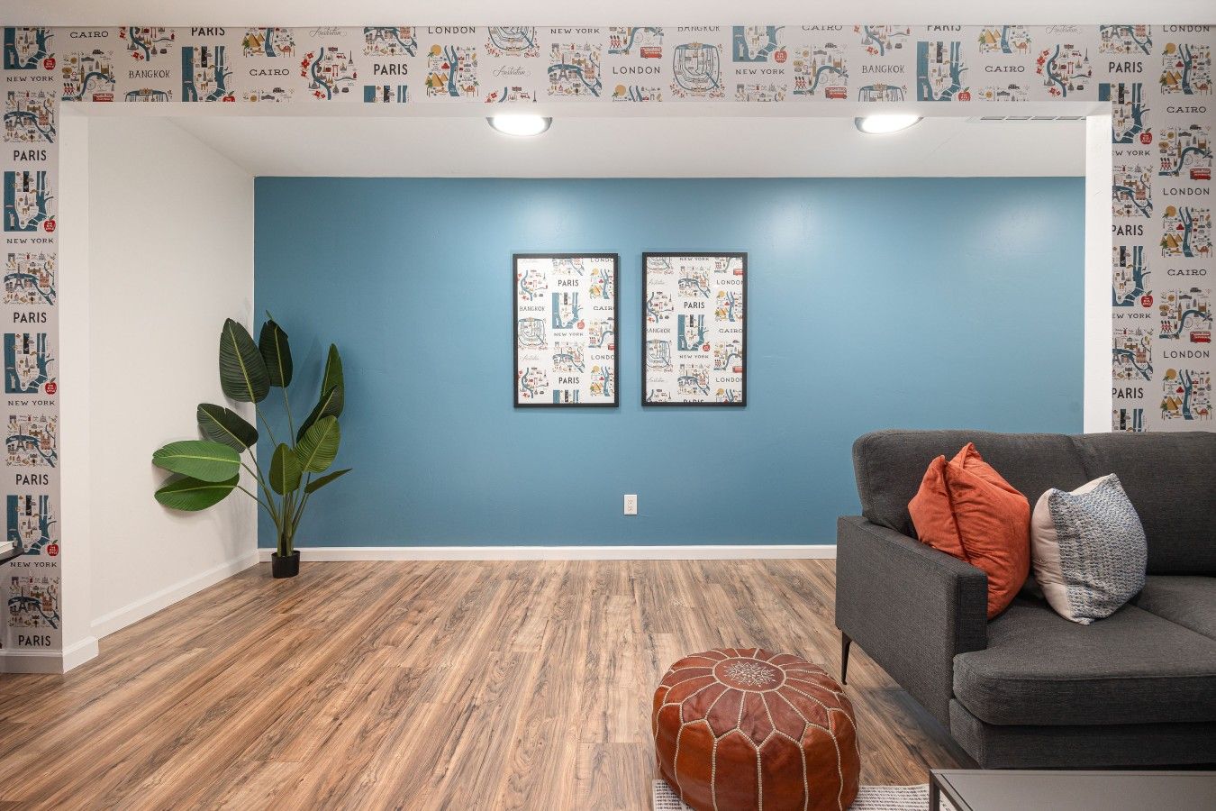 Une pièce avec un mur bleu, deux affiches encadrées représentant des paysages urbains, un canapé gris avec des coussins, une grande plante verte et un plancher en bois.