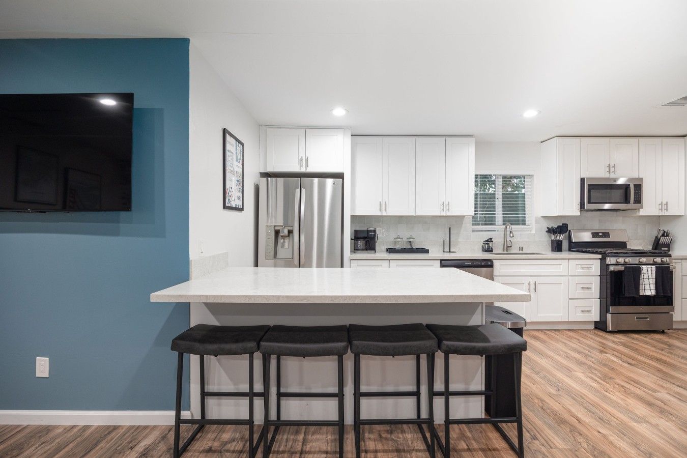 A modern kitchen with white cabinets, a stainless steel refrigerator, a microwave, and a stove. There are three bar stools at the island.