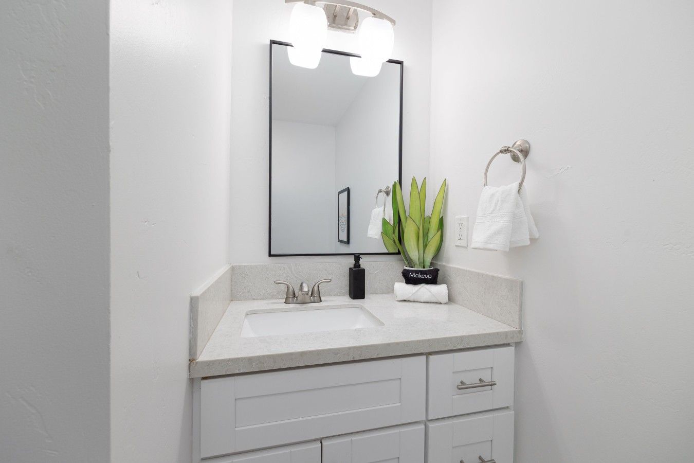 A bathroom with a white sink, a mirror with lights, a potted plant, and a towel rack.