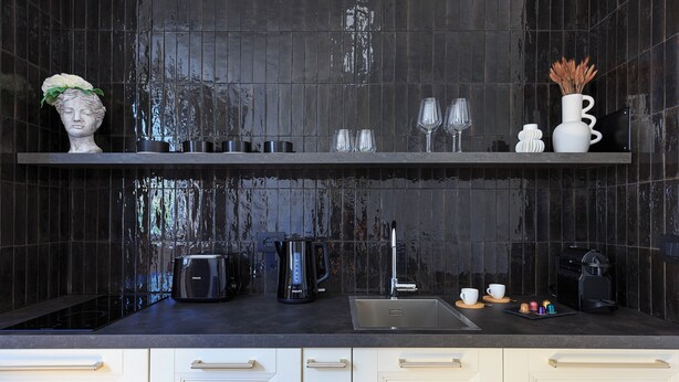 Et moderne køkken med en backsplash af sorte fliser, en stålvask, kaffemaskine og elkedel på bordpladen.