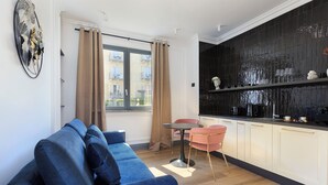 En moderne stue med en blå sofa, et lille rundt bord og pink farvede stole. Der er et køkken med sorte fliser og hvide skabe.