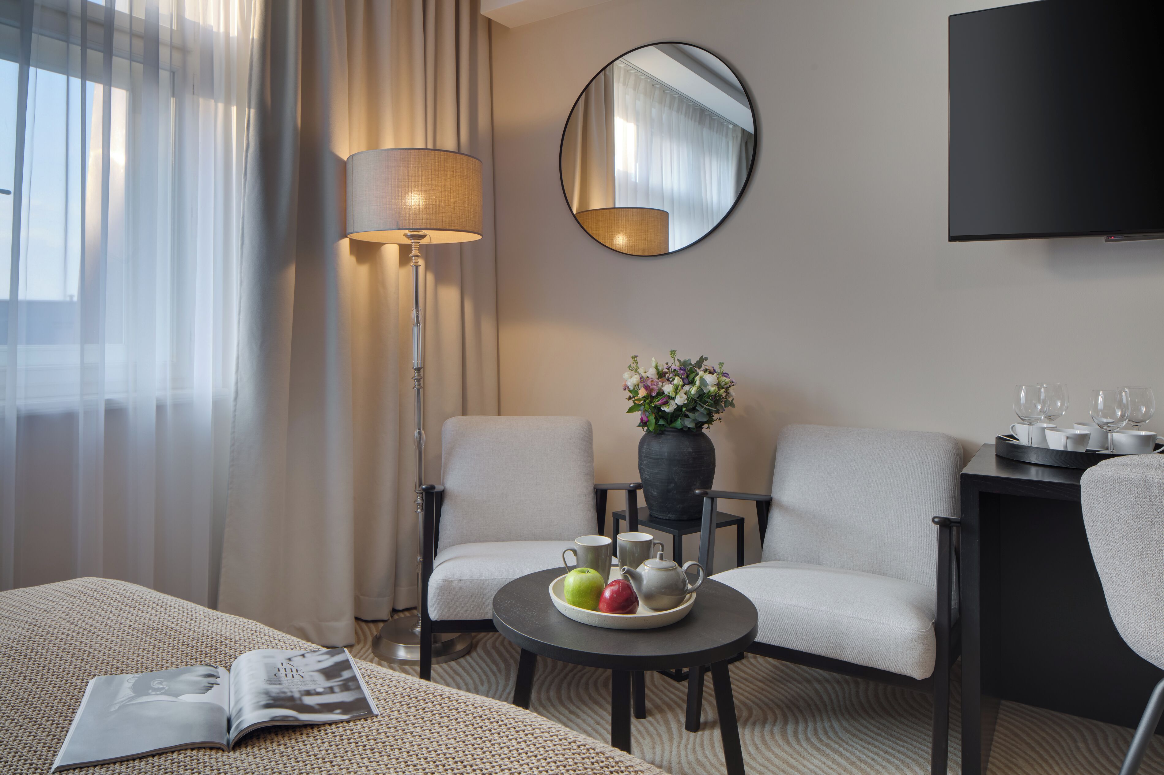 Habitación de hotel con cama, dos sillones, una mesita con frutero, un jarrón con flores, una lámpara, un espejo y un televisor de pantalla plana.