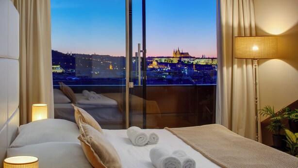 Ein Hotelzimmer mit einem Bett, zwei aufgerollten Handtüchern, einer Nachttischlampe, einem Fenster mit Stadtblick und einer Topfpflanze.