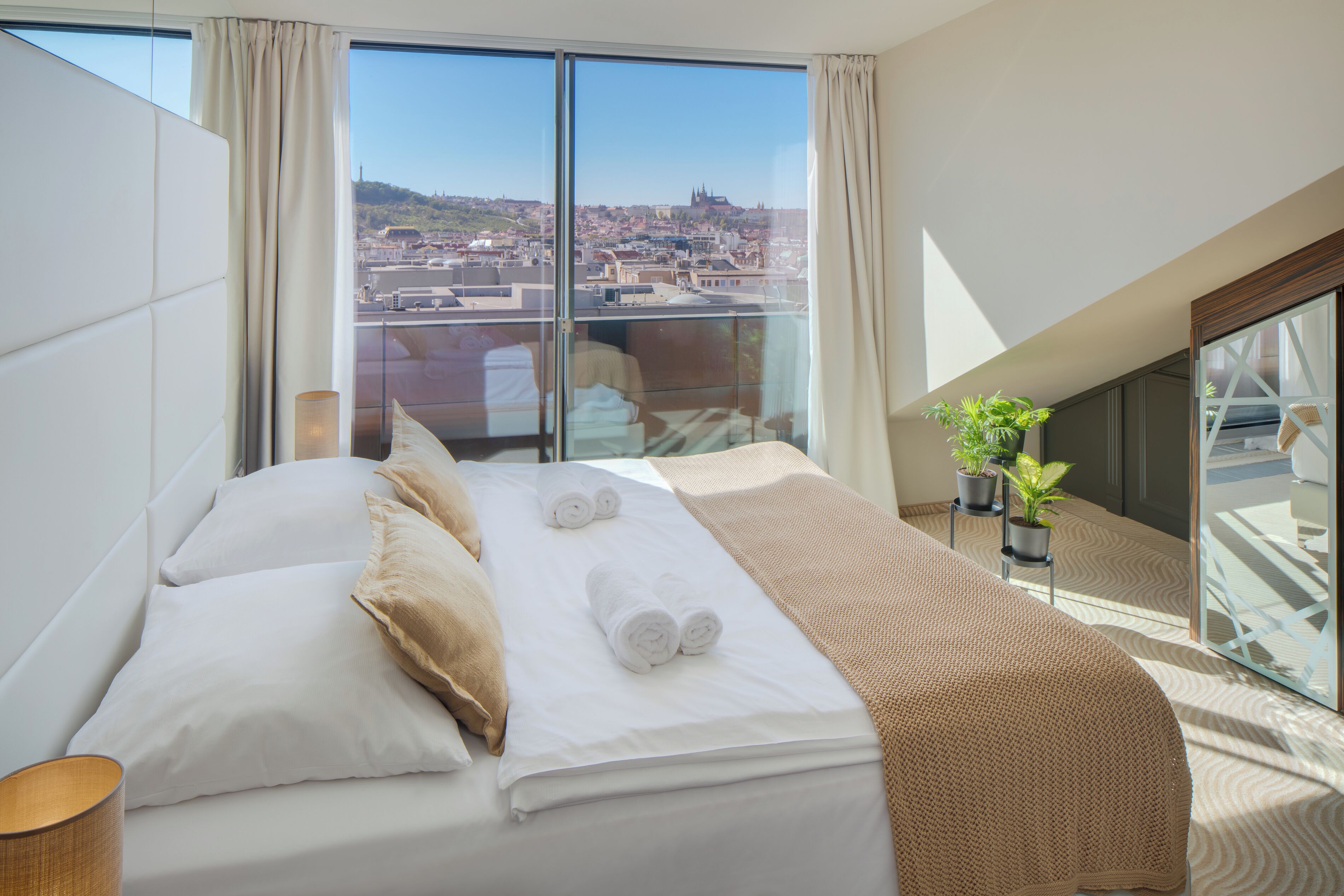 A hotel room with a large bed, a view of a cityscape, and a balcony with potted plants.