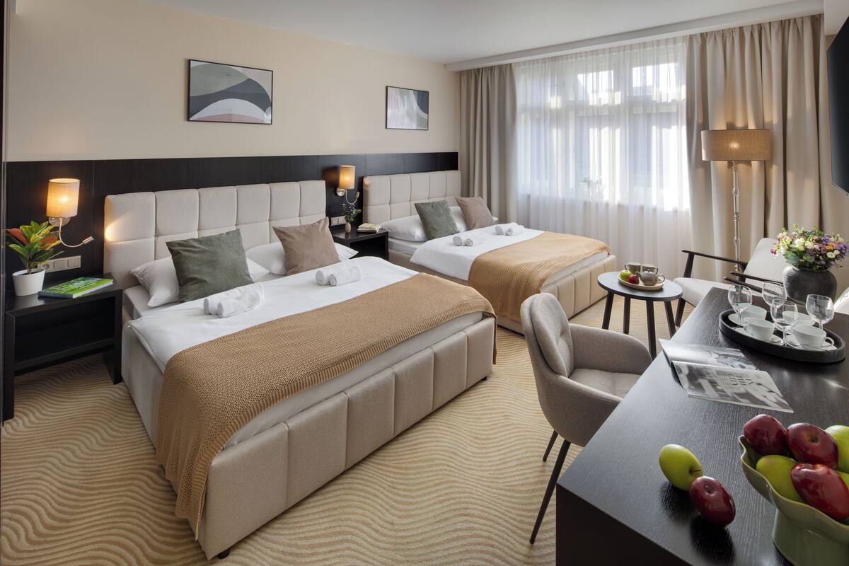 A hotel room with two beds, a dining table with a tray of fruit, a lamp, and a view of the outdoors.