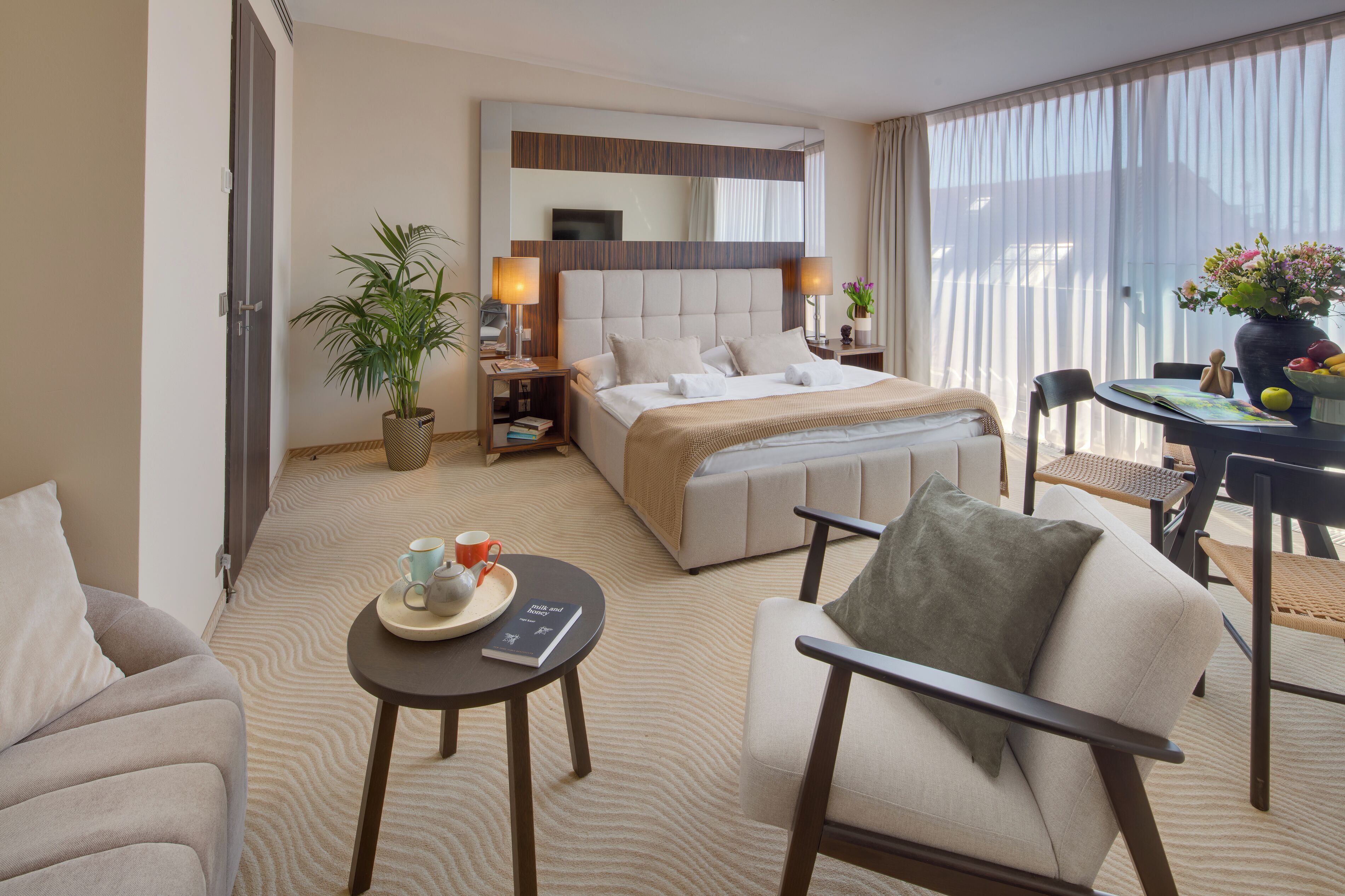 A hotel room with a bed, a sofa, a dining table with chairs, a small side table, a potted plant, and a large window with curtains.