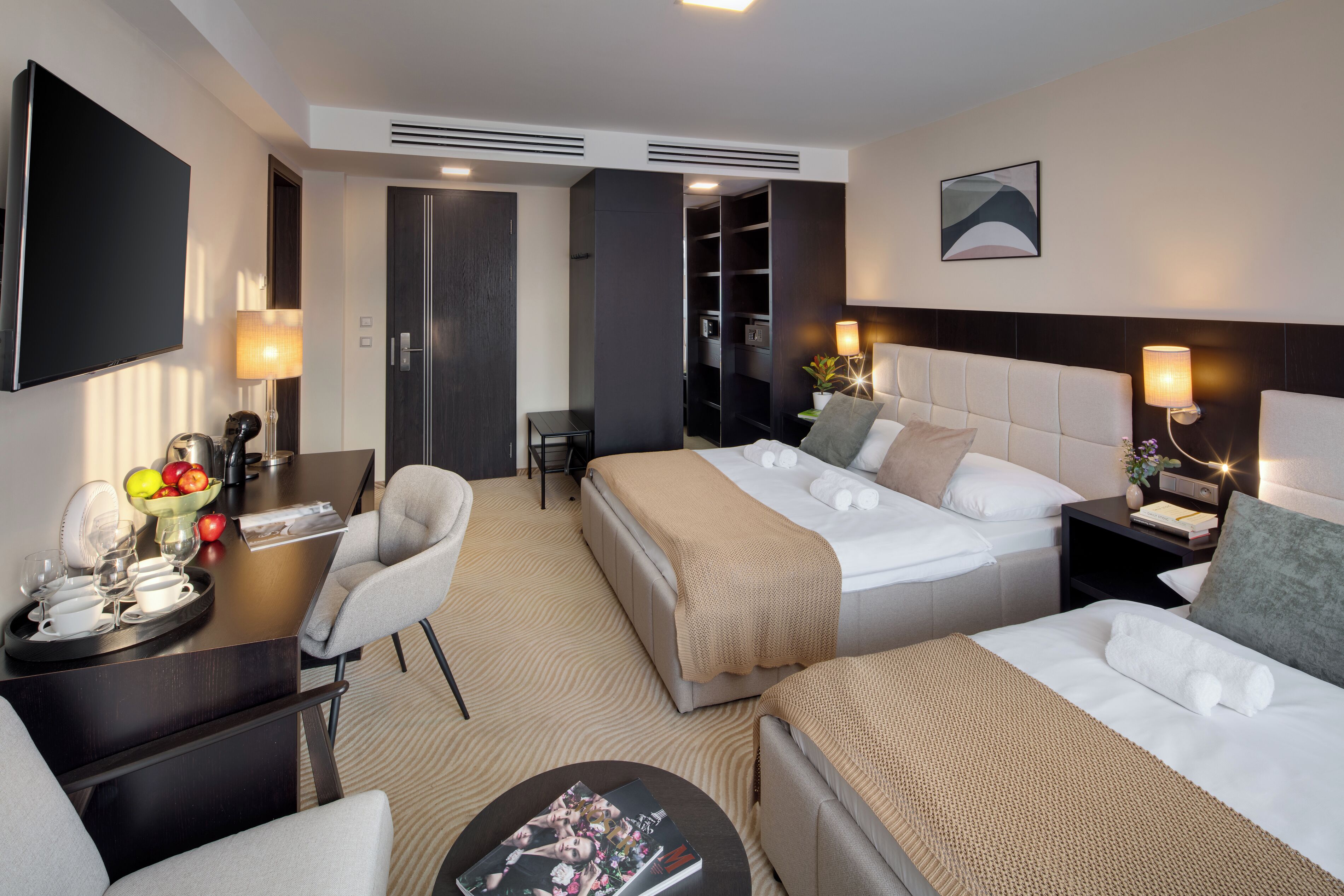 A hotel room with a TV, two beds, a desk with a tray of fruit, and a seating area.