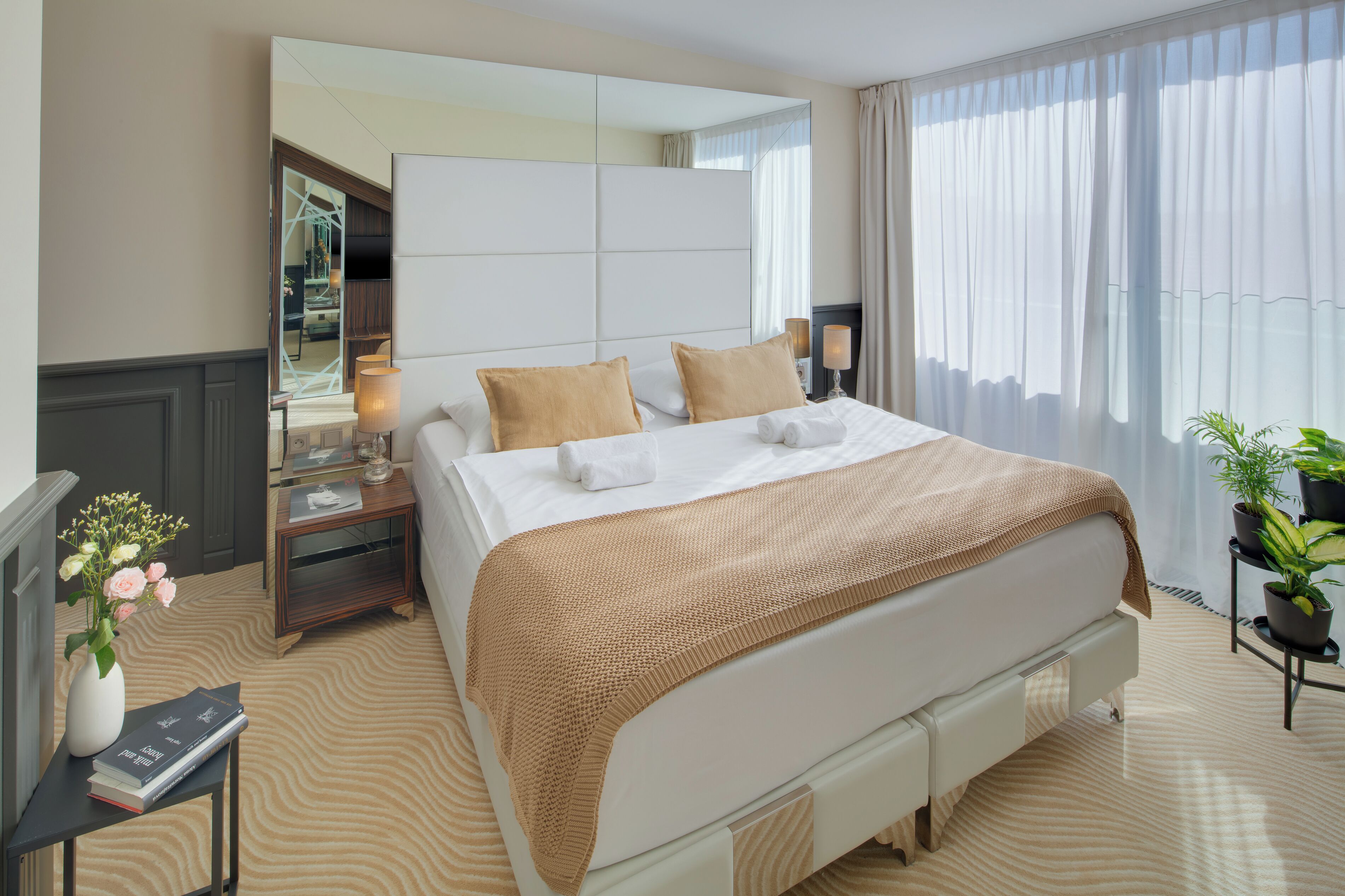 A hotel room with a large bed, bedside tables, a mirror, a potted plant, and a view of the outdoors.