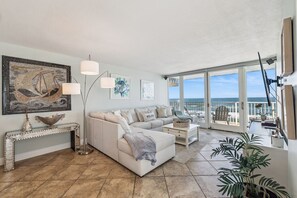 A spacious living room with a large window overlooking the sea, a white sofa, a console table, and a floor lamp. - Southeast Serenity-Luxury Oceanfront Condo | New Smyrna Beach 1 Bedroom | 1 Bathroom (New Smyrna Beach)
