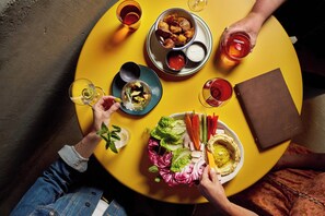A round yellow table with various food items and drinks, surrounded by people. - All Inn (Denver)