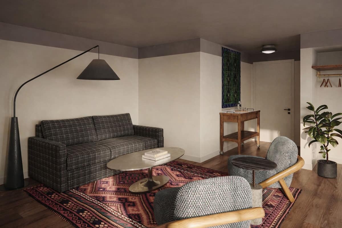 Una sala de estar con un sofá, una mesa de centro, una silla, una lámpara de pie, una mesita con una planta y una alfombra estampada.