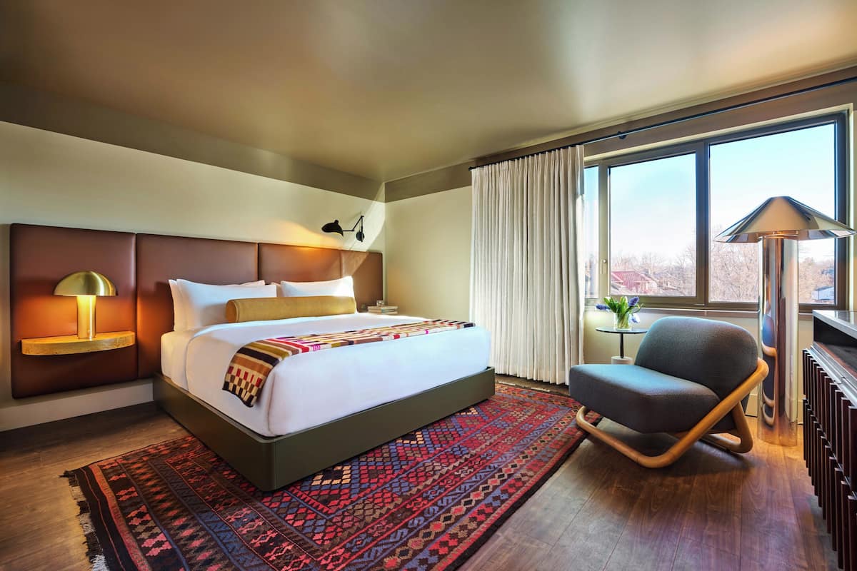 Una habitación de hotel con una cama grande, una mesita de noche con lámpara, una silla, una ventana con cortinas y un radiador.