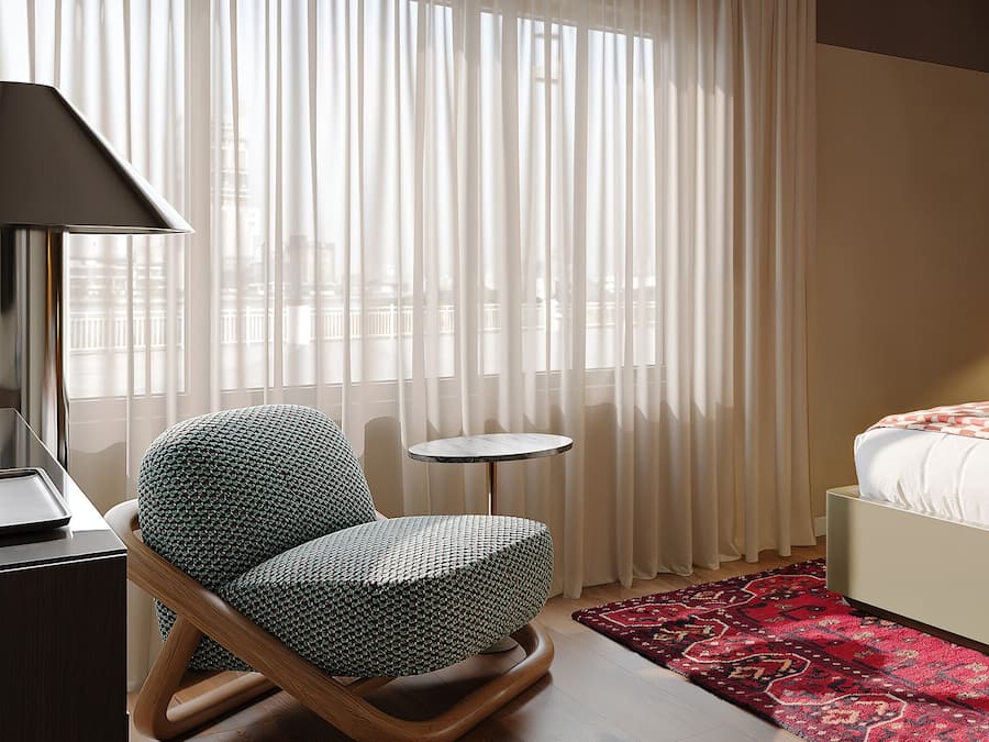 A hotel room with a bed, a chair, a small table, a lamp, and a window with curtains.