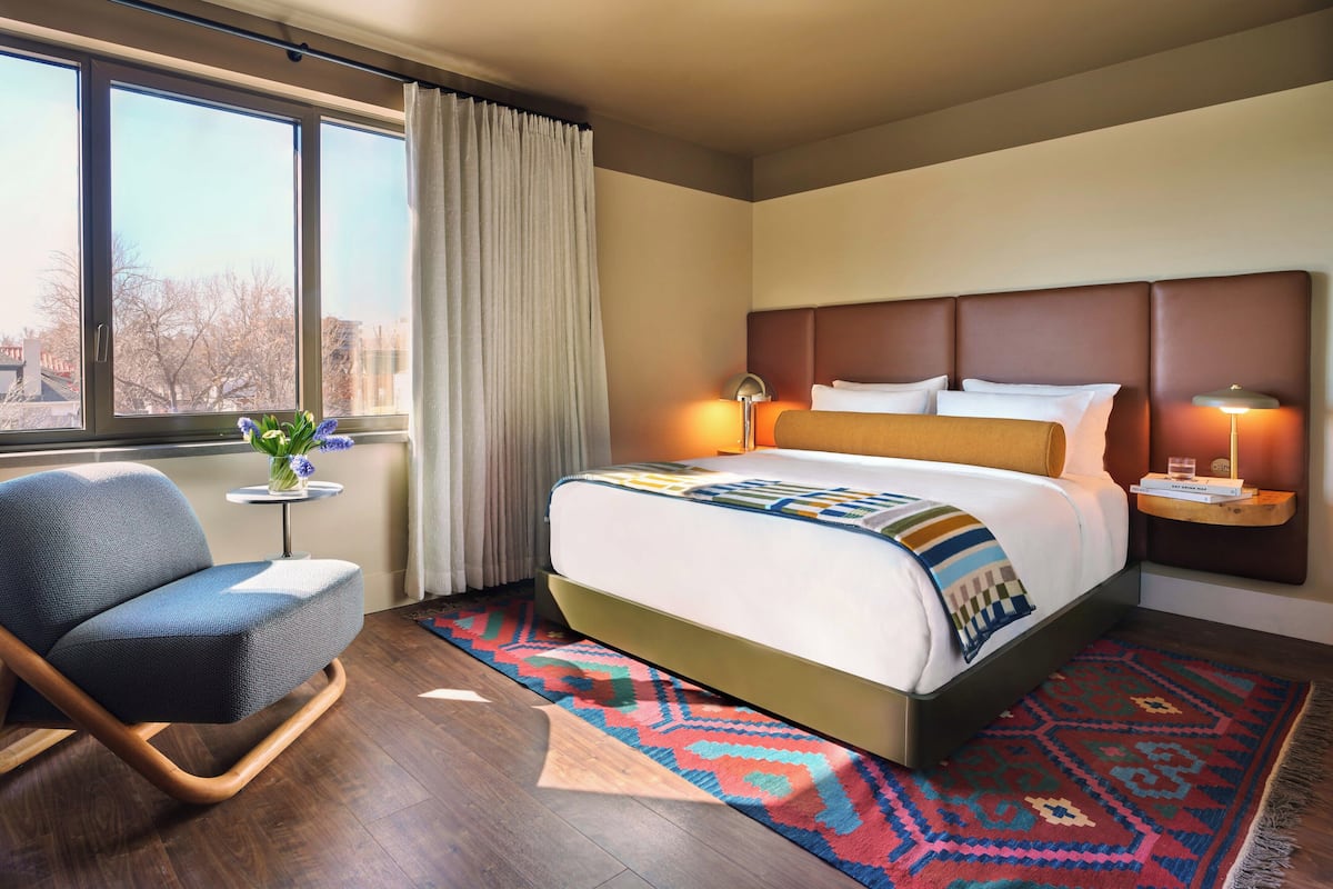 Habitación de hotel con cama, una silla, una mesita con un florero y vistas al exterior.