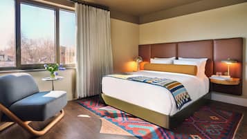 Habitación de hotel con cama, una silla, una mesita con un florero y vistas al exterior.