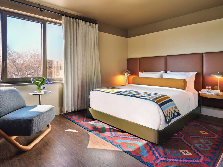 Habitación de hotel con cama, una silla, una mesita con un florero y vistas al exterior.