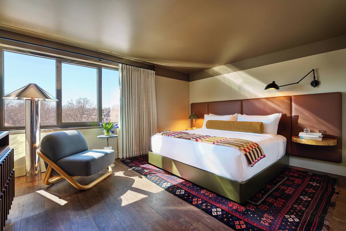 Habitación de hotel moderna con una cama grande, una silla, una lámpara y vistas al exterior.