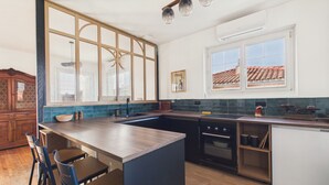 A modern kitchen with a large island, wooden chairs, a built-in oven, and a window with a view of the roof. - Comfortable house for 8 with garden (Saint-Gilles-Croix-de-Vie)