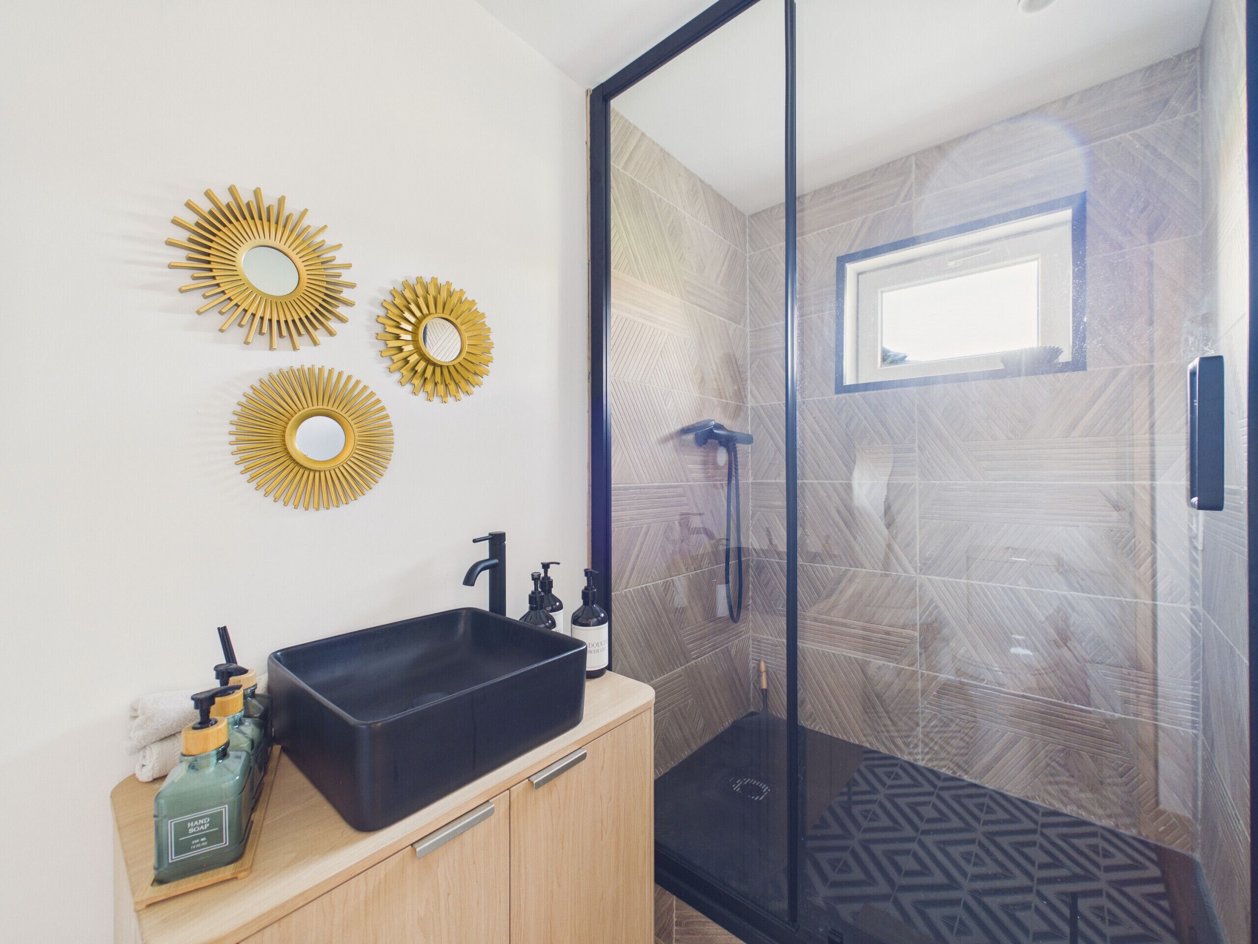 Une salle de bain moderne avec un lavabo noir, trois miroirs dorés en forme de soleil et une douche dont le sol est orné d’un motif géométrique.