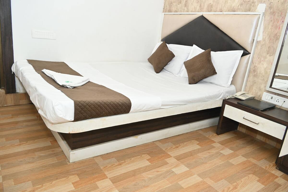 Un lit simple avec un cadre en bois, une literie blanche et des oreillers marron. Il y a une table de chevet avec un téléphone et une prise électrique.