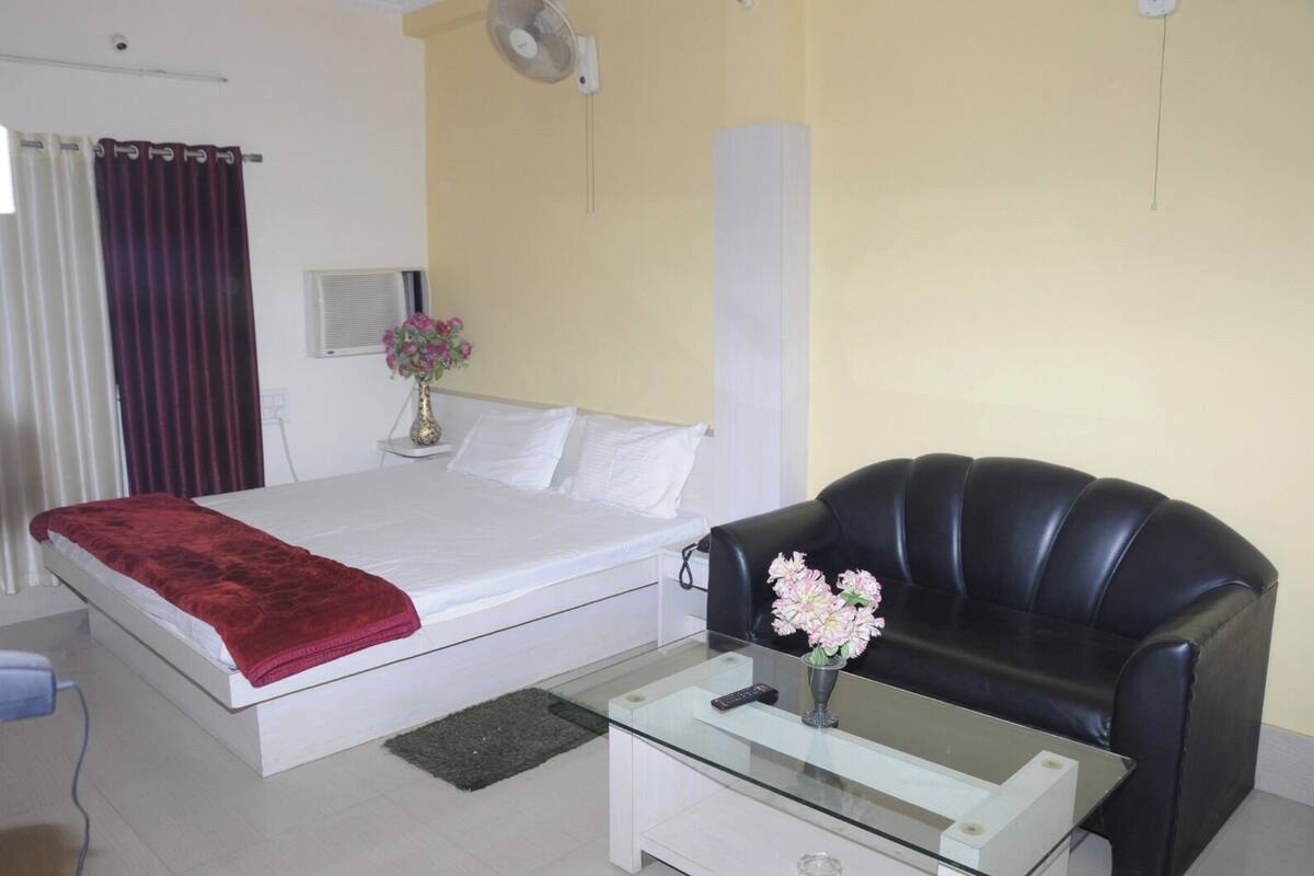Une chambre d’hôtel avec un lit, un canapé en cuir noir, une table basse en verre, un vase avec des fleurs, un climatiseur fixé au mur et une fenêtre avec des rideaux.