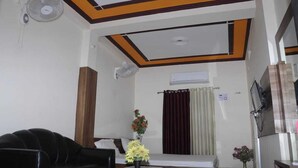 Een woonkamer met een zwarte leren bank, een glazen salontafel en een vaas met bloemen. Er is een plafondventilator, een airconditioningsysteem en een raam met gordijnen.