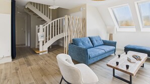 A modern living room with a blue sofa, a white armchair, and a wooden coffee table. There is a staircase with a white railing leading to another room. - 100m from the beach – House for 8 with garden (Fouesnant)