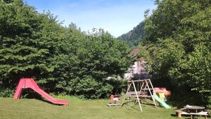 Un espace extérieur verdoyant avec un toboggan rouge, une balançoire en bois et des tables de pique-nique.