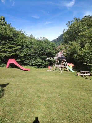 Un espacio al aire libre con césped, un tobogán rojo, un conjunto de columpios de madera y mesas de picnic.