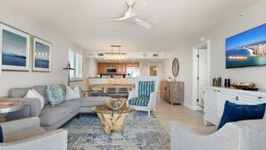 A spacious living room with a grey sofa, a white armchair, and a coffee table. A kitchen is visible in the background, and a TV is mounted on the wall. - Beachfront 3BR, 2BA Apollo condo with pool, WiFi, and heated Gulf views. (Marco Island)