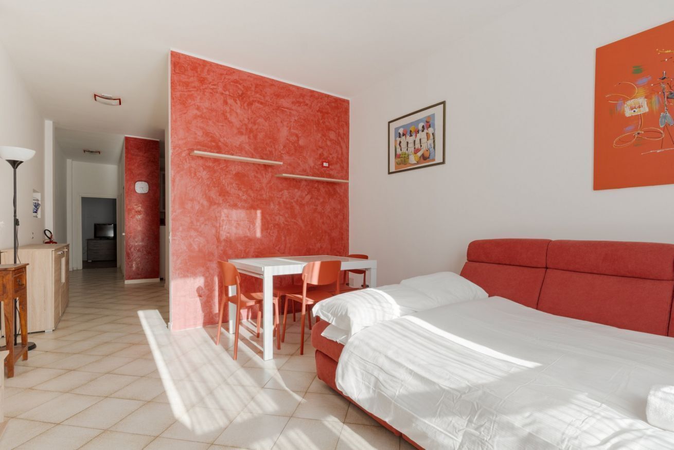 A bedroom with a bed, a desk, and a chair, a red accent wall, and a painting on the wall.