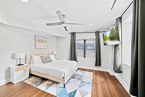 A modern hotel room with a bed, bedside tables, a ceiling fan, a window with curtains, and a view of a river. - Prime 3BR Philly Pad Fits 6 Guests Walk Everywhere (Philadelphia)