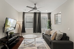 A modern living room with a flat-screen TV, a ceiling fan, a sofa, a coffee table, and a large window with curtains. - Stylish 3BR Sleeps 6 Oasis | Near The Liberty Bell (Philadelphia)