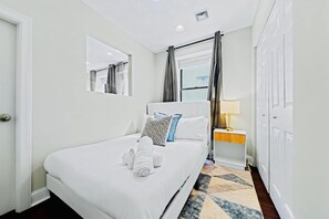 A hotel room with a bed, a bedside table with a lamp, a mirror, and a window with curtains. - Prime 3BR Philly Pad Fits 6 Guests Walk Everywhere (Philadelphia)