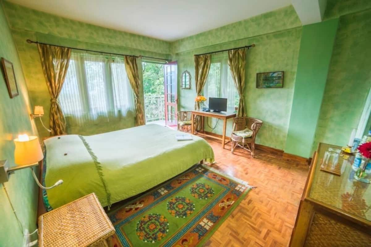 A room with a bed, a desk with a computer, a chair, and a small table with a lamp and flowers.