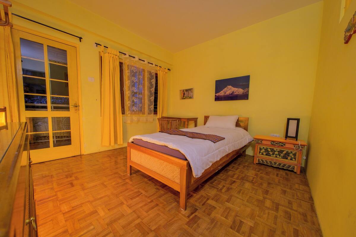 A bedroom with a wooden bed, a nightstand, a dresser, a window with curtains, and a door with a view.